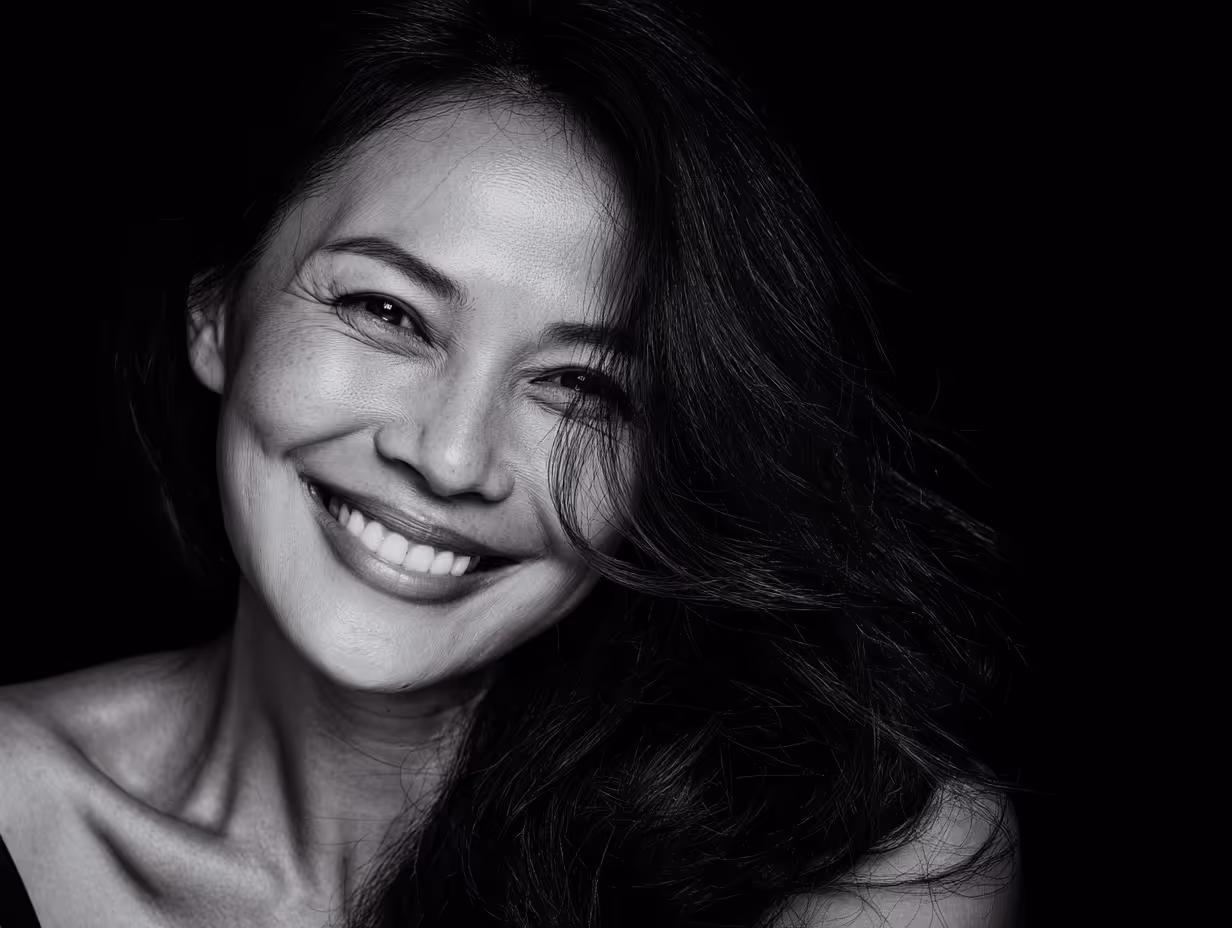 Black and white close-up portrait of a smiling woman with long dark hair against a black background.