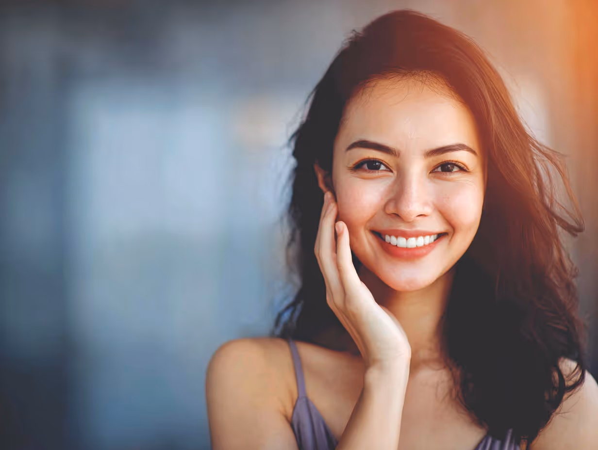 Young woman with dark hair smiling brightly and resting her hand on her cheek against a blurred background.